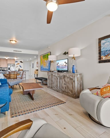 Open-plan living room with blue sofa, white armchairs, wooden ceiling fan, and beach-themed artwork on walls