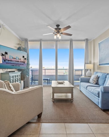 Beachfront condo living room with blue sofa, tan armchair, ocean view from balcony doors, and ceiling fan