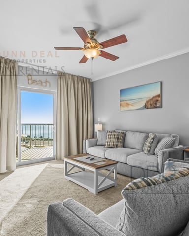 Beachfront condo living room with gray sofas, ceiling fan, balcony view, and beach artwork