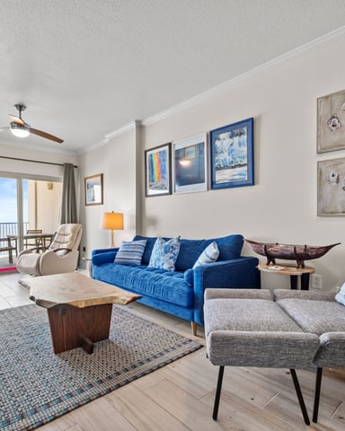 Bright beachfront living room with blue sofa, gray armchair, ocean view balcony, and coastal wall art