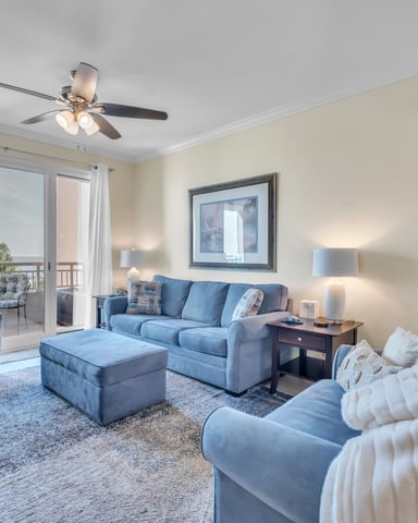Spacious living room with blue upholstered seating, ceiling fan, and glass doors opening to a balcony with water views