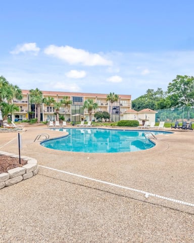 Resort pool with concrete deck, palm trees, lounge chairs, and multi-story beige buildings with red roof