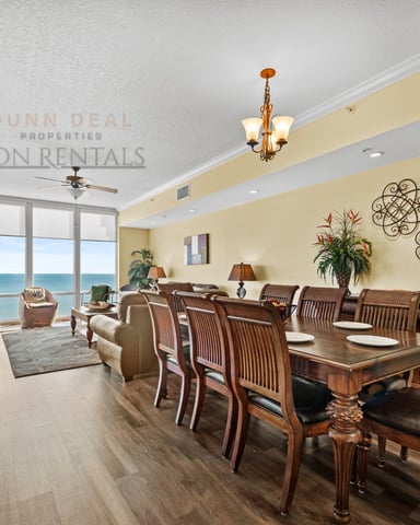 Open-plan dining and living room with cream walls, wooden dining table, ocean view through floor-to-ceiling windows, and coastal decor.