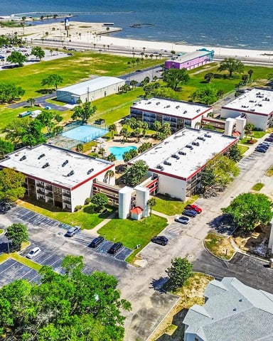 Aerial view of beachfront resort complex with pools, parking lots, green lawns, and high-rise hotels overlooking turquoise water
