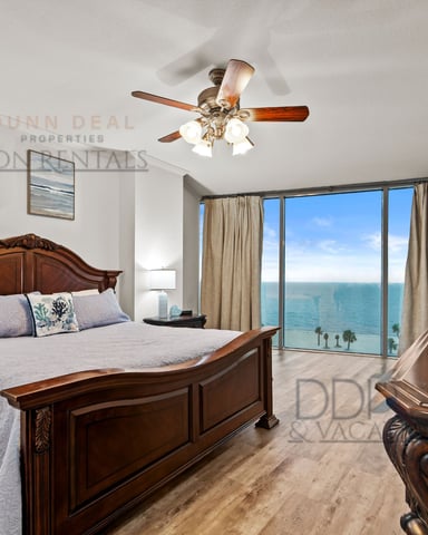 Bedroom with wooden bed, ceiling fan, and floor-to-ceiling windows overlooking ocean and palm trees