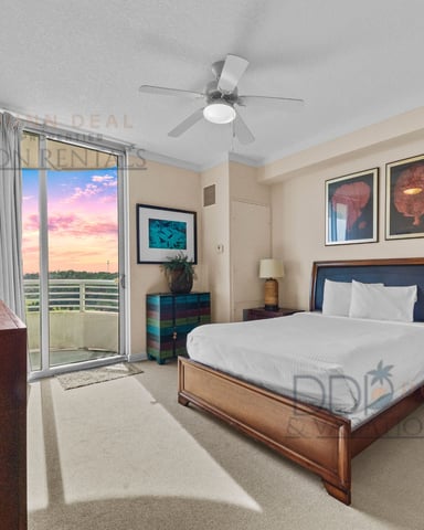 Hotel bedroom with queen bed, desk, balcony overlooking sunset and water, ceiling fan, and framed artwork on walls