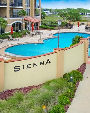 Resort pool area with turquoise swimming pools, tan stucco buildings, palm trees, and red umbrellas