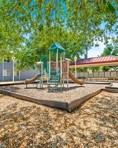 Playground with green slide structure and sandbox surrounded by trees, residential buildings and covered patio in background