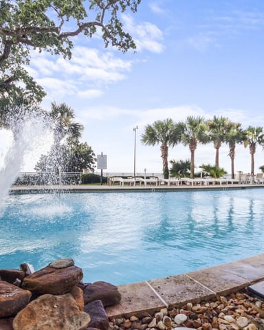 Resort pool with fountain, palm trees, and waterfront lounge area under blue sky