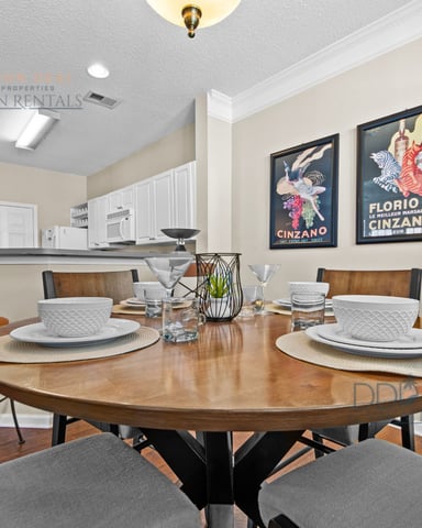 Open-plan dining and kitchen area with round wood table, place settings, vintage beverage posters, and white cabinetry