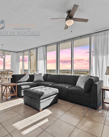 Open-plan living room with black sectional sofa, floor-to-ceiling windows overlooking pink sunset ocean view, ceiling fan, and dining area.