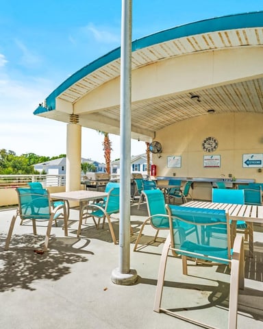 Covered poolside pavilion with turquoise lounge chairs, tables, and cream-colored walls under blue metal roof