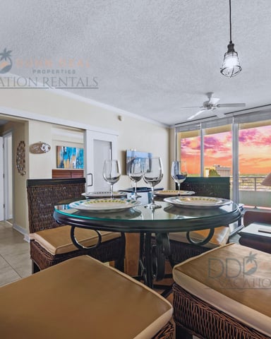 Dining room with round glass table, wicker chairs, and floor-to-ceiling windows overlooking sunset ocean view