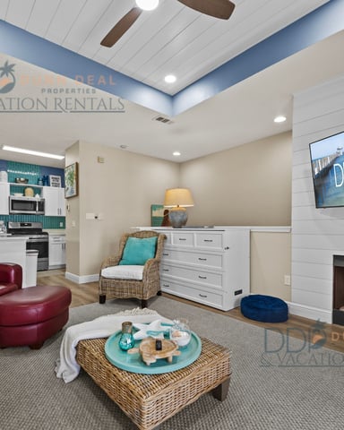 Open-plan living room with burgundy leather sofa, fireplace, kitchen with teal backsplash, and flat-screen TV displaying ocean view.