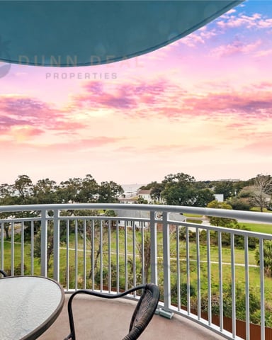 Curved balcony with metal railing overlooking coastal landscape at sunset, with pink and orange sky, green fields, and distant water