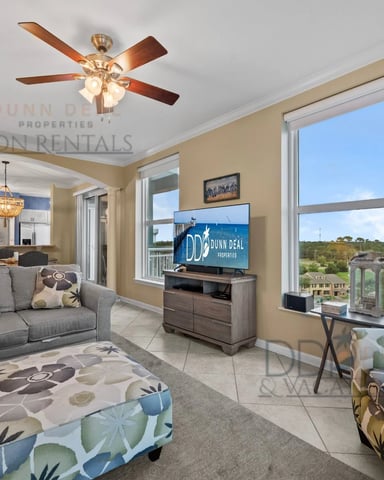Open-plan living room with gray sofa, patterned ottomans, TV, ceiling fan, and large windows overlooking coastal landscape