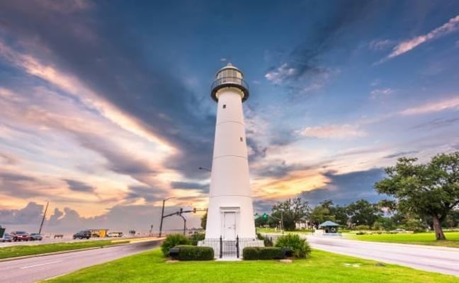White lighthouse with lantern room on green lawn at sunset, surrounded by trees and parked cars